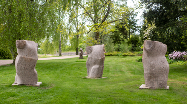 Tre stenskulpturer med formen liknande kroppar. Står i gräset med träd i bakgrunden. 