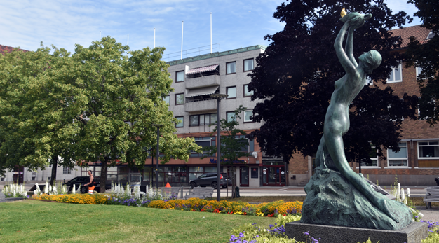 Skulptur av en kvinna som håller upp en brinnande låga. Skulpturen är placerad i en park med planteringar och träd. 