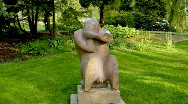 Granitskulptur av en man som håller en hand för ena ögat. Bakgrunden består av grönska, träd och buskar