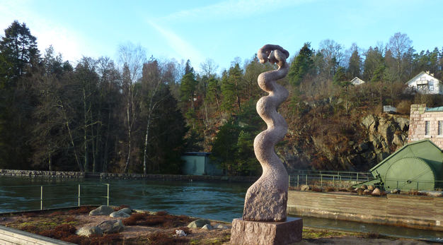 Granitskulptur formad som en skruv placerad på en bro med vatten i bakgrunden.