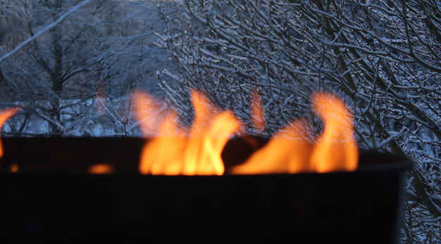 Lågor i en grill. I bakgrunden är det träd med snö på grenarna. 