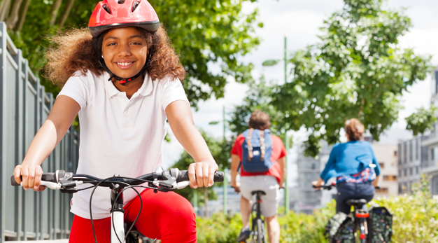 En glad flicka med röd cykelhjälm cyklar i en stad i solen. 