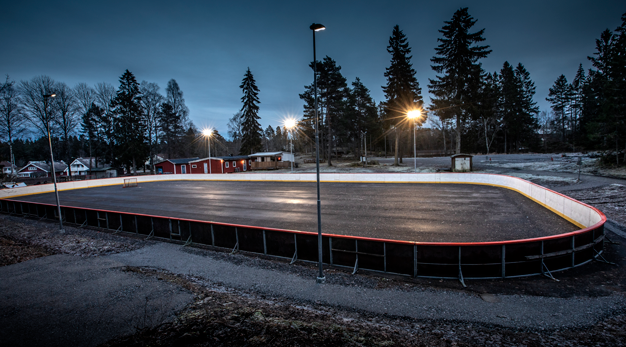 En asfalterad yta med en hockeyrink runt. Två mål står på ytan. Det är skymningsljus och lamporna runt rinken är tända. 