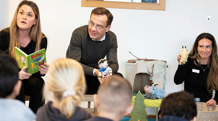 Tre vuxna sitter framför en barngrupp. Man ser barnens ryggar och inte deras ansikten. Till vänster sitter en kvinnlig förskollärare och håller en bok med titeln "Petter och hans fyra getter". I mitten sitter statsminister Ulf Kristersson och håller ett litet gosedjur. Till höger sitter en kvinlig förskollärare. 