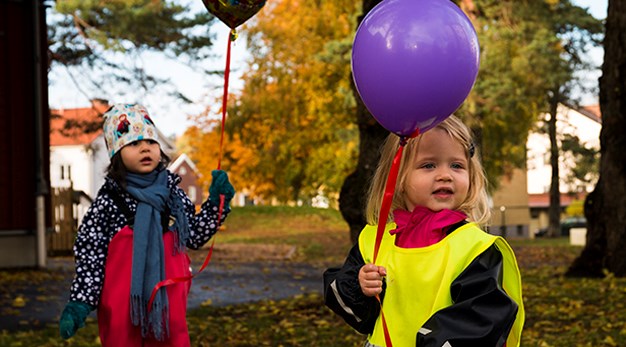 Bilden föreställer två små flickor iklädda regnkläder och reflexväst som håller i varsin ballong. Ballongerna är lila. I bakgrunden syns orangea och röda träd. Fotografiet är taget på hösten. Marken är full med löv. 