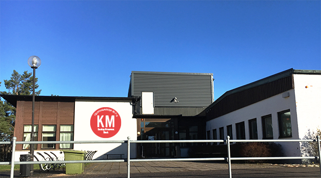 Bilden föreställer Slättbergsskolans huvudentré. Byggnaden är vit i grunden och har bruna partier i trä. Framför skolans entré finns det cykelställ och bakom skolan syns en klarblå himmel. 