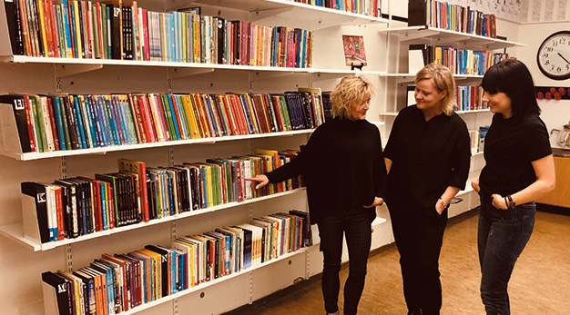 Bilden föreställer tre kvinnor, alla klädda i svart, som står framför en vägg fylld med böcker. Kvinnorna står på rad snett i bild och kvinnan längst till vänster pekar på en bok. De andra kvinnorna tittar nyfiket på boken.
