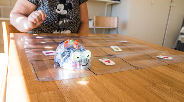 Fotografiet föreställer en liten robot som ser ut som en mus. Roboten kör runt på ett duk som är placerad på ett bord i trä. I bakgrunden syns en kvinna. Kvinnan har en tröja med Musse Pigg på.