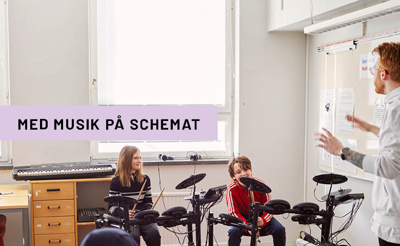 Tjej och kille som spelar trummor i lektionssal med lärare som instruerar. 