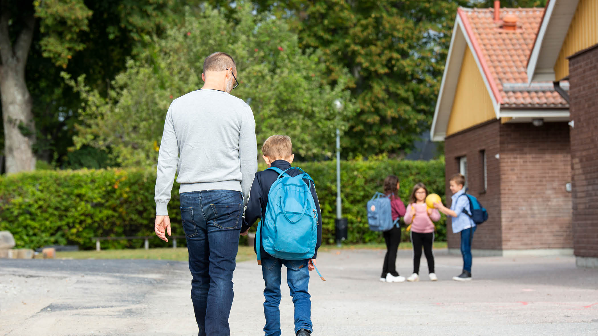 Pappa som håller barn med ryggsäck i handen på väg mot skolan. I bakgrunden syns tre barn.