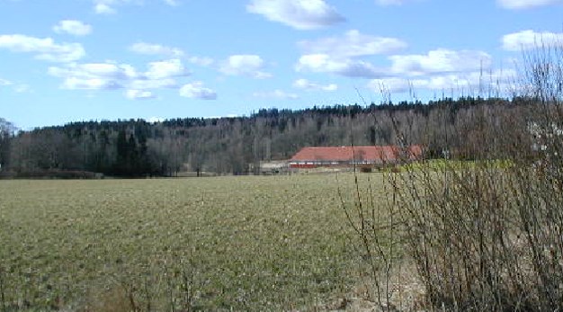 Solig dag med blå himmel, åkermark med en röd lada i bakgrunden. 