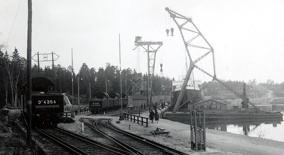 Svartvit historisk bild på rysskranen där man även kan se ett fartyg och två tåg på spåren som då gick genom området. 
